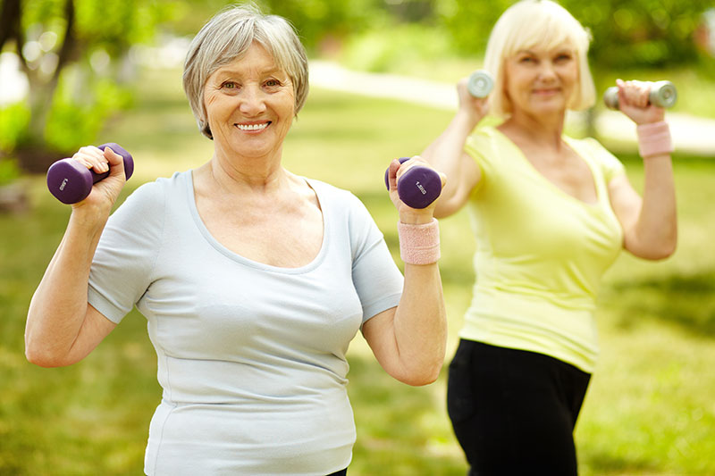 Two older women exercising with dumbbells in a park.
